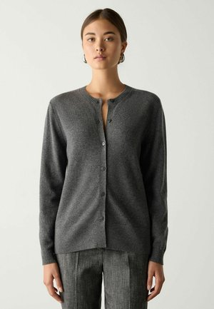 Junge Frau mit dunklem Haar in einem niedrigen Dutt trägt eine geknöpfte graue Strickjacke und graue Hose vor einem schlichten hellen Hintergrund.