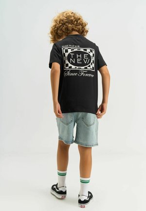 Barn med krøllet hår iført en sort t-shirt med teksten "Make Way for The New Since Forever", lyse denimshorts, hvide sokker og sorte sneakers.