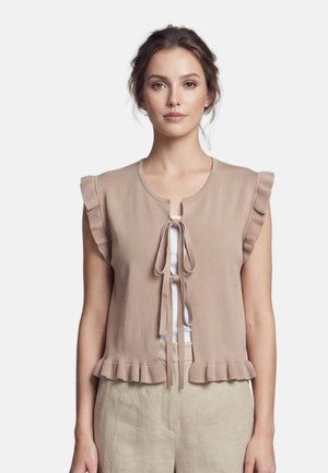 Vrouw met bruin haar draagt een mouwloos beige gebreid vest met ruches en striksluitingen over een wit topje, gecombineerd met lichtbeige broek.