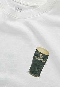 Biała koszulka z nadrukiem szklanki piwa Guinness z pianką na wierzchu oraz logo Guinness na przodzie.