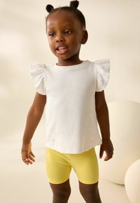 T-shirt bianco a coste con maniche corte a volant e pantaloni gialli. L'outfit presenta un design semplice, una texture morbida e colori vivaci.