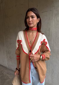 Nő bézs és fehér kardigánban, piros szegéllyel, piros nyakkendővel, arany ékszerekkel, világoskék farmernadrágban, bézs borítéktáskával a kezében, egy betonfal mellett áll.