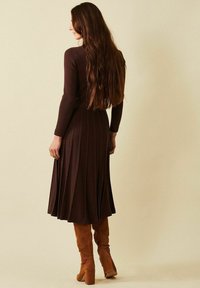 Nainen, jolla on pitkät ruskeat hiukset, pukeutunut tummanruskeaan ribbimäiseen pitkähihaiseen mekkoon ja polvimittaisiin vaaleanruskeisiin mokkasaappaisiin, seisoo yksinkertaista beigeä taustaa vasten.