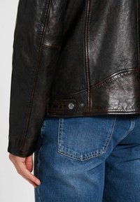 Braune Lederjacke mit strukturierter Oberfläche, sichtbaren Nähten und Druckknopfverschluss an der Taille. Getragen über blauen Jeans.