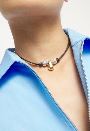 Gros plan sur un cou portant un collier en corde sombre avec des perles en argent et un pendentif abstrait doré, au-dessus d'une chemise bleu clair partiellement zippée.