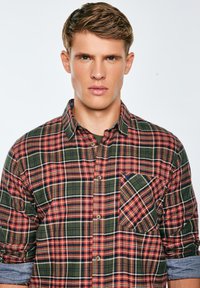 Camasa în carouri roșie, verde și bleumarin, cu guler căptușit, mâneci lungi cu manșete răsfrânte și un buzunar pe piept cu un model diagonal.