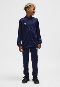 Navy tracksuit made of smooth fabric, featuring a zip-up jacket with a logo and pants with black chevron stripes along the sides.