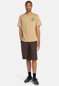 Beige t-shirt med et lille grafisk motiv på brystet, parret med mørkebrune shorts. Båret med sorte sportssko og sokker.