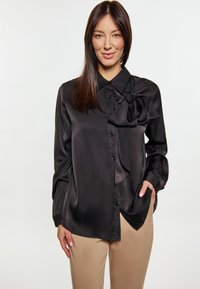 Camicia nera in satin con un grande fiocco dettagliato sul collo, maniche lunghe e texture liscia. Abbinata a pantaloni beige.
