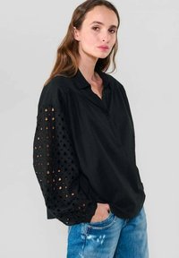 Frau mit langen braunen Haaren, die eine schwarze Bluse mit durchbrochenen Puffärmeln und blaue, verwaschene Jeans trägt, steht mit einer Hand in der Tasche.