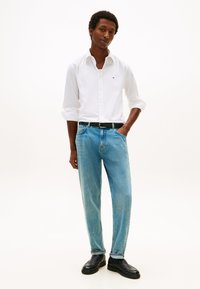 Ung mann som står med den ene hånden i bukselommen, iført en hvit skjorte med knapper, lyseblå jeans, svart belte og svarte sko mot hvit bakgrunn.