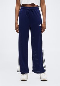 Άτομο που φοράει αθλητικό παντελόνι με φαρδιές μπλε σκούρες γραμμές και λευκές ρίγες στα πλάγια, καθώς και λευκά αθλητικά παπούτσια, στέκεται μπροστά σε ένα απλό φόντο.