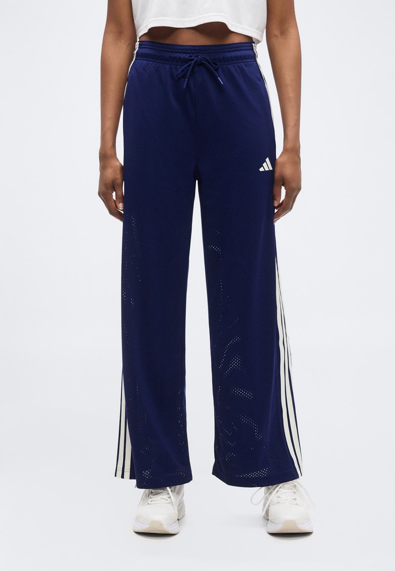 Άτομο που φοράει αθλητικό παντελόνι με φαρδιές μπλε σκούρες γραμμές και λευκές ρίγες στα πλάγια, καθώς και λευκά αθλητικά παπούτσια, στέκεται μπροστά σε ένα απλό φόντο.