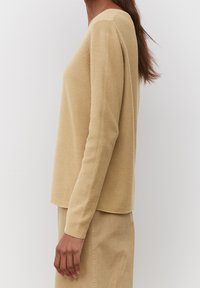 Beige neulepaita, jossa pitkät hihat, pyöreä pääntie ja resorikuvio, yhdistetty vaaleanruskeaan hameeseen. Sivukuva, joka näyttää istuvuuden ja kankaan.