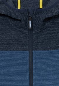 Modrá fleecová bunda s predným zipsom a textúrovanou tkaninou. Výrazné farebne rozdelené časti v tmavej a strednej modrej, doplnené žltou vnútornou podšívkou.