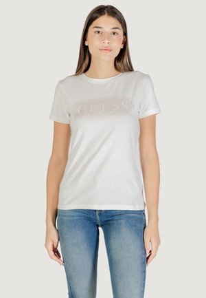 Camiseta blanca de algodón con mangas enrolladas, que presenta un diseño del logo "GUESS" brillante en el frontal. Combinada con unos jeans de mezclilla azules.