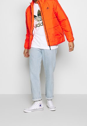 Osoba ubrana w jasnopomarańczową kurtkę puchową Adidas, białą koszulkę z logo Adidas, jasnoniebieskie podwinięte dżinsy oraz białe wysokie trampki Converse.