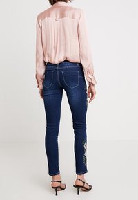 Slim-fit mörkblå jeans med broderad blommönster på vänster ben, kombinerat med en ljusrosa satinblus med lång ärm och stiletto-klackar.
