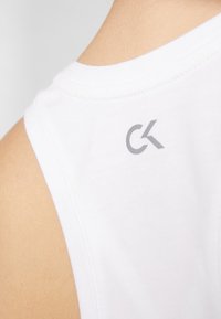 Valkoinen hihaton puuvillapaitapusero, jossa on pieni harmaa "CK"-logo takana. Sileä pinta ja pyöreä pääntie siisteine ompeleineen.