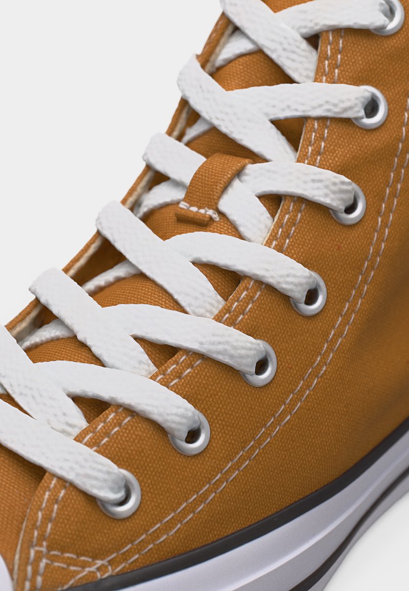 Zapatilla deportiva de lona marrón con cordones blancos entrecruzados pasados por ojales de metal y suela de goma blanca visible.