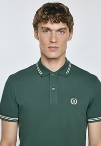 Polo tričko v tmavě zelené barvě, s dvoubarevnými knoflíky, pruhovaným límečkem a rukávy, s bílým logem na levé hrudi.
