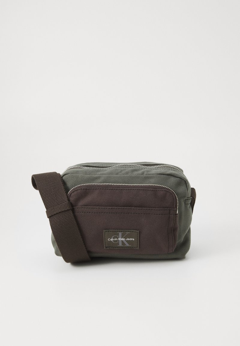 Zaļa un brūna auduma crossbody soma ar rāvējslēdzēju, regulējamu siksnu un Calvin Klein logo uzšuvē priekšpusē. Kompakta taisnstūra forma.