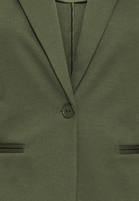 Blazer verde oliva con tessuto testurizzato, chiusura con un bottone singolo e due tasche frontali con dettagli neri. Vestibilità sartoriale con revers affilati.