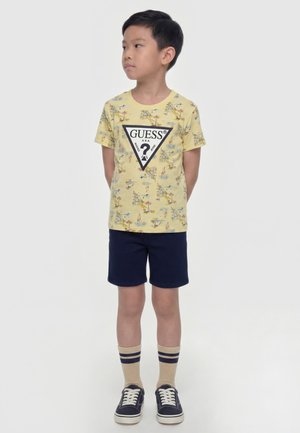 Ragazzo con una t-shirt gialla a motivi Guess, pantaloncini blu navy, calzini beige a righe e sneakers blu navy, in piedi con le mani dietro la schiena che guarda a sinistra.