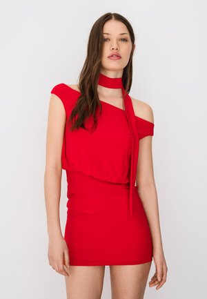 Femme aux longs cheveux bruns portant une robe courte rouge à épaules dénudées avec un collier attaché, debout devant un fond blanc uni.