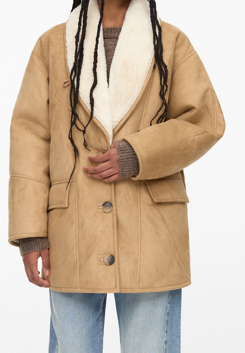 Personne portant un manteau en peau retournée beige avec un grand col et des boutons, par-dessus un pull en maille marron et un jean bleu clair.