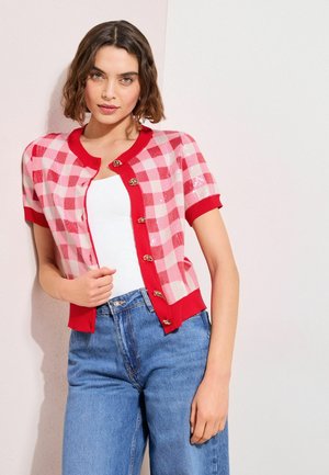 Femme portant un cardigan à manches courtes à carreaux rouges et roses par-dessus un haut blanc avec un jean taille haute bleu, debout contre un mur bicolore.