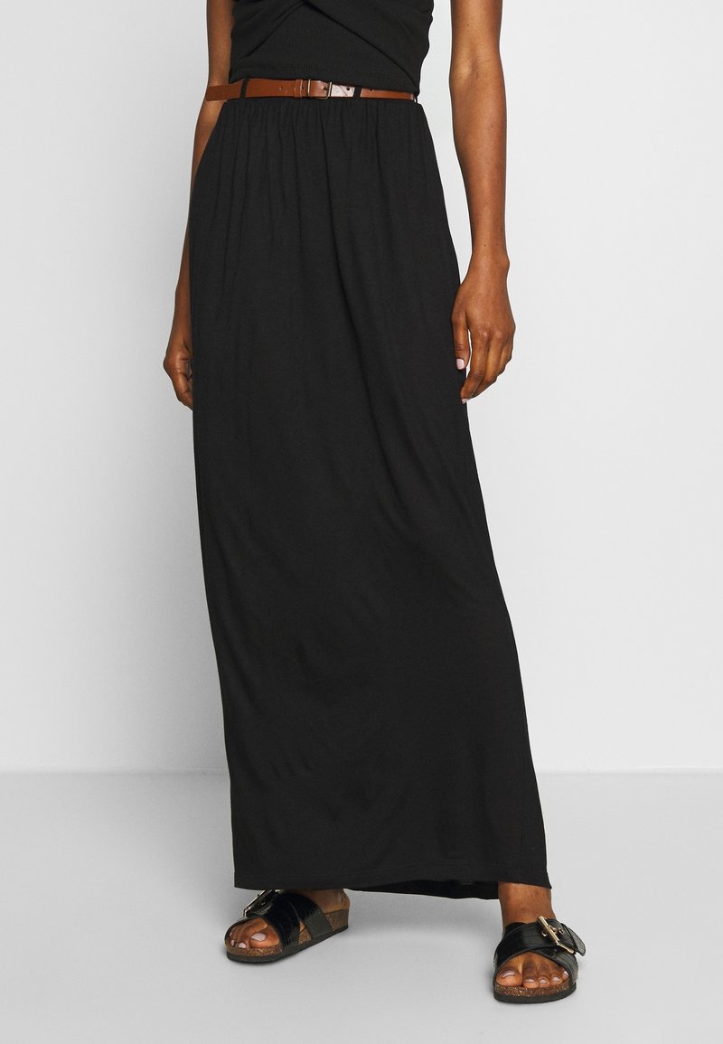 Falda maxi negra con cintura elástica, combinada con un cinturón marrón; usada con sandalias marrones que tienen una hebilla decorativa. Textura suave.