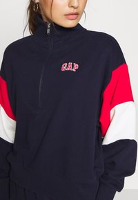 Felpa con zip di colore blu navy con maniche a righe rosse e bianche. Presenta il logo "GAP" ricamato sul petto. Tessuto morbido e vestibilità comoda.