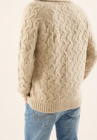 Beiger, kabelstrick gestrickter Pullover mit geripptem Saum und Bündchen, versehen mit einem strukturierten Muster; kombiniert mit hellblauen Jeans, zeigt die Rückansicht.