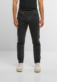 Jeans in denim nero con una vestibilità slim, dotati di cinque tasche e un dettaglio con zip rossa. Indossati con scarpe da ginnastica chiare.