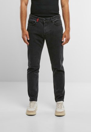 Vaqueros de mezclilla negra con corte slim, que cuentan con cinco bolsillos y detalle de tirador de cremallera roja. Combinados con zapatillas de color claro.