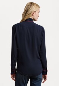 Cămașă albastră cu mânecă lungă, cu cusături contrastante roșii, manșete cu nasturi și o textură fină. Dispune de un guler clasic și un tiv curbat.
