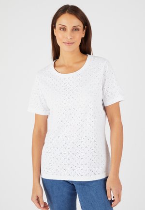 Femme aux longs cheveux bruns portant un top blanc à manches courtes en œillet et un jean bleu, debout devant un fond clair uni.