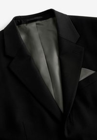 Veste de costume noire avec doublure intérieure grise et pochette grise assortie dans la poche poitrine, présentée sur fond blanc.