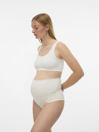 Bodoča mati v belih materinskih spodnjih perilih, stoji v profilu ob enobarvni beli ozadju, z rokami za hrbtom.