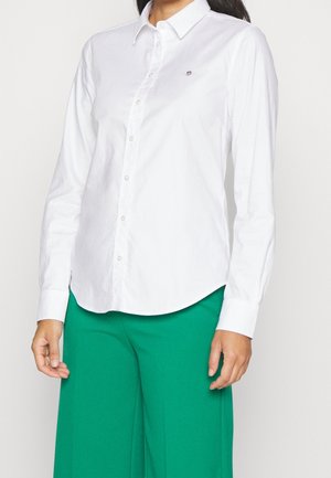 Femme portant une chemise blanche à manches longues boutonnée et un pantalon vert taille haute à jambes larges sur un fond uni.