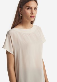 Ung kvinna med långt brunt hår som bär en ljusbeige kortärmad blus och guldfärgade ringformade örhängen, vänd framåt mot en vit bakgrund.