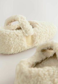 Mjuka krämfärgade tofflor gjorda av texturerat tyg med en fluffig, pälsliknande yta, med en rem och ett meshningsöppning för stöd.