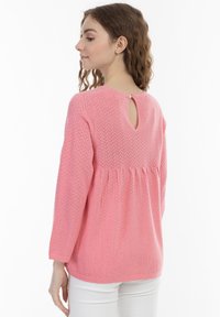 Maglione pullover rosa chiaro, con texture, dettagli a chiave sul retro, maniche 3/4 e vita arricciata. Abbinato a pantaloni bianchi.