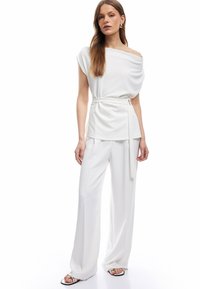 Top bianco a spalle scoperte con tessuto drappeggiato e pantaloni a palazzo coordinati. L'outfit presenta una cintura, una texture liscia e un design semplice.