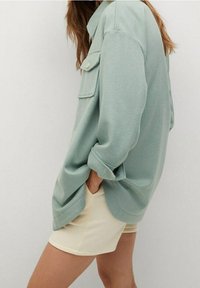 Camicia oversize verde chiaro con colletto e dettagli sulla tasca, abbinata a pantaloni corti beige. I tessuti sembrano morbidi e rilassati.