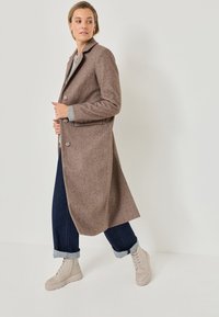 Brauner Wollmischungsmantel mit Reverskragen, Frontknöpfen, seitlichen Taschen und langen Ärmeln, kombiniert mit hochgekrempelten dunklen Jeans und hellen Stiefeln.