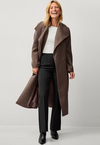 Cappotto lungo in misto lana marrone con ampi rever, doppi bottoni e una fodera interna liscia, abbinato a pantaloni neri e stivali alla caviglia.