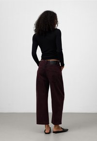 Pantaloni a gamba larga bordeaux con vita alta, tasche posteriori visibili e una patch in pelle sulla vita. Il modello indossa una maglietta nera e sandali.