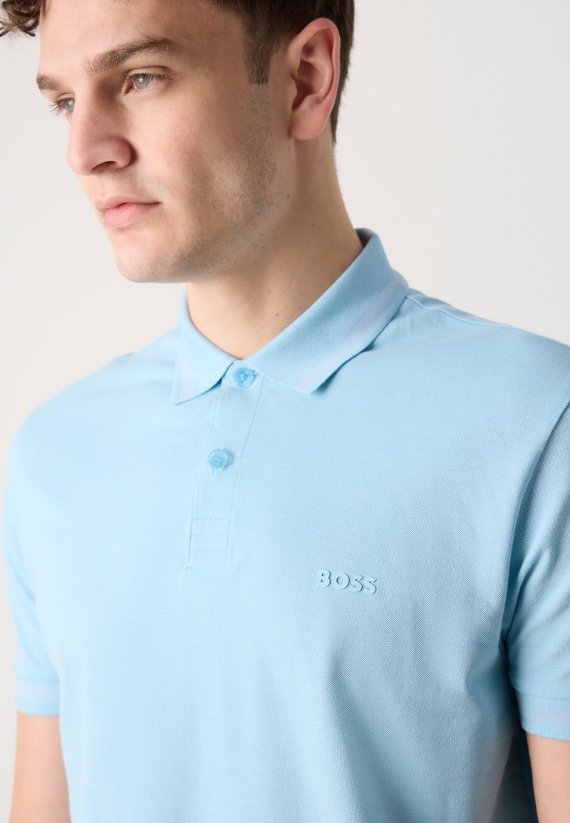 Lichtblauwe polo in strakke stof, met een klassieke kraag, twee knopen en een reliëf logo op de borst.